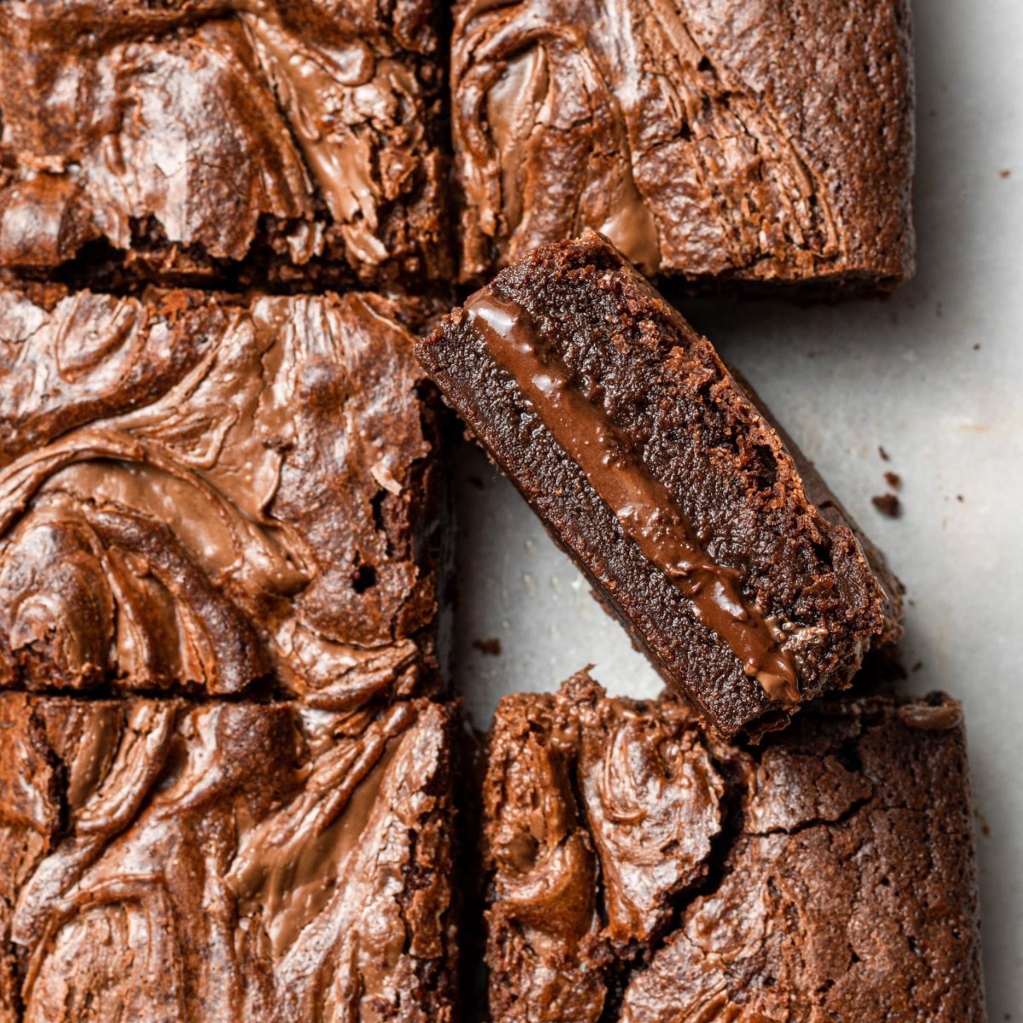 Several chocolate brownies arranged on a flat surface, with one turned on its side to showcase a thick Nutella layer between fudgy brownie layers. The marbled top adds visual richness and texture.