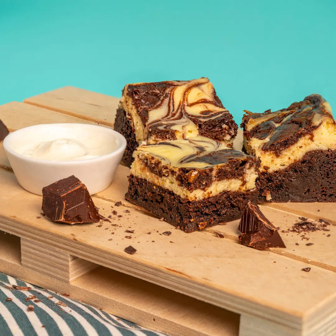 Three marble Cheesecake Chaos brownies with rich chocolate and creamy cheesecake swirls placed on a small wooden pallet, styled with a scoop of cream in a white bowl and scattered dark chocolate chunks against a bright turquoise background. The Brownie Girl dessert photography
