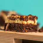 A close-up of a Cheesecake Chaos brownie featuring a fudgy chocolate base, a smooth cheesecake centre, and a marbled chocolate top, displayed on a wooden surface with a turquoise backdrop. The Brownie Girl product image