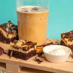 A glass of iced coffee with condensation paired with several marbled Cheesecake Chaos brownies topped with chocolate chunks, served beside a bowl of whipped cream on a wooden surface with a turquoise background. The Brownie Girl dessert pairing