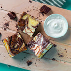 Three marble Cheesecake Chaos brownies with bold chocolate and cheesecake swirls, surrounded by chocolate shavings and a bowl of cream cheese on a light wooden surface with a striped cloth in the background. The Brownie Girl product photography