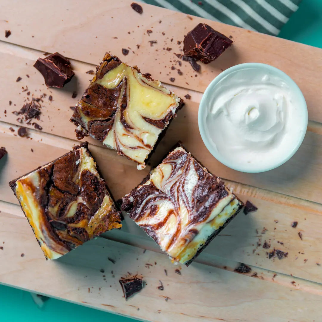 Three marble Cheesecake Chaos brownies with bold chocolate and cheesecake swirls, surrounded by chocolate shavings and a bowl of cream cheese on a light wooden surface with a striped cloth in the background. The Brownie Girl product photography