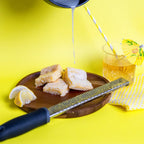 A hand pouring white icing over Lemon Kiss Blondie's on a wooden plate, with lemon wedges, a zester, and a glass of iced citrus drink with a yellow umbrella in the background, all set on a bold yellow surface. The Brownie Girl summer dessert scene.