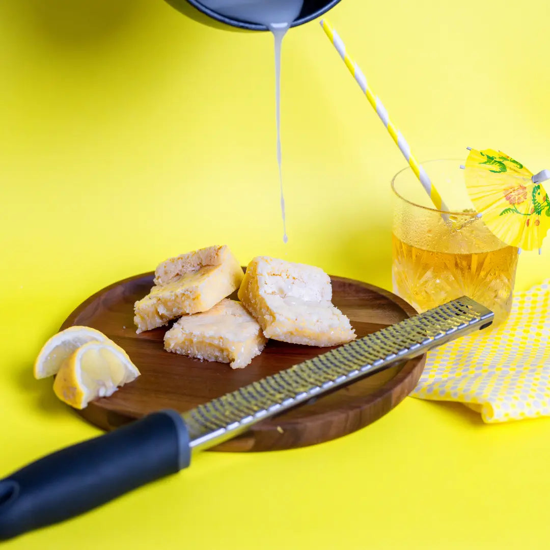 A hand pouring white icing over Lemon Kiss Blondie's on a wooden plate, with lemon wedges, a zester, and a glass of iced citrus drink with a yellow umbrella in the background, all set on a bold yellow surface. The Brownie Girl summer dessert scene.