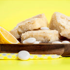 Several Lemon Kiss Blondie's on a wooden tray with scattered white chocolate pieces and a lemon wedge, styled against a vibrant yellow background. The Brownie Girl citrus themed product image