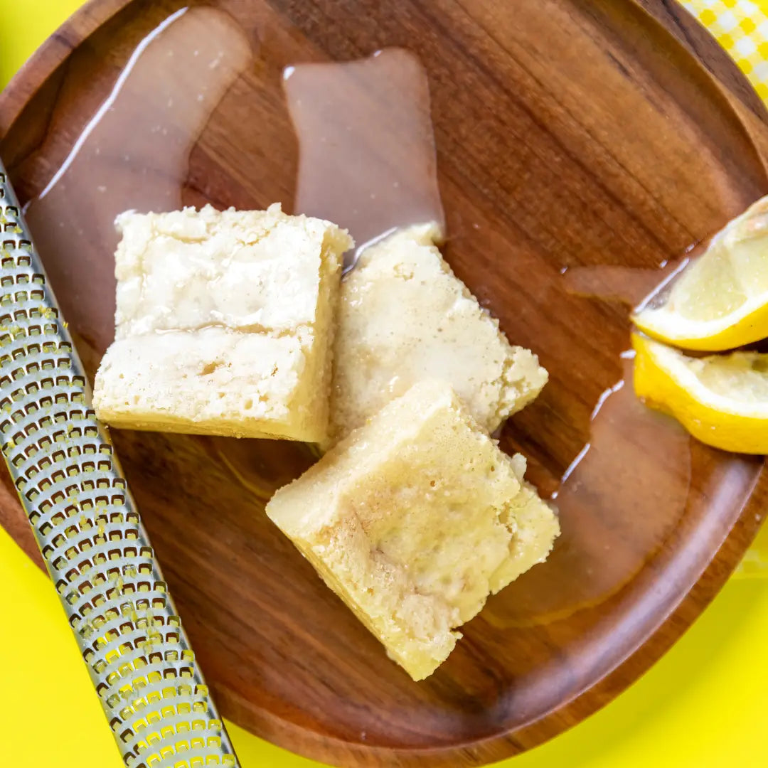 Three Lemon Kiss Blondie's on a round wooden plate drizzled with glossy glaze, placed beside lemon wedges and a metal zester on a bright yellow backdrop. The Brownie Girl fresh citrus dessert styling