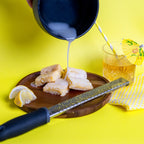 Lemon Kiss Blondie's on a wooden plate with white icing being poured from above, surrounded by lemon wedges, a zester, and a glass of iced citrus drink on a bright yellow background. The Brownie Girl summer dessert photography