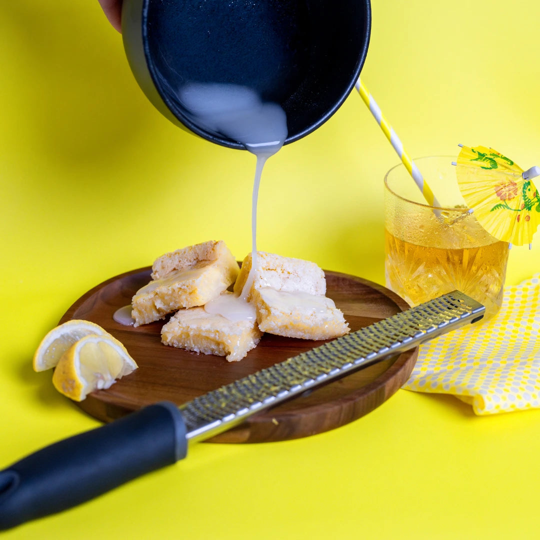 Lemon Kiss Blondie's on a wooden plate with white icing being poured from above, surrounded by lemon wedges, a zester, and a glass of iced citrus drink on a bright yellow background. The Brownie Girl summer dessert photography