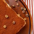 A close-up of the Pick‑Me‑Up tiramisu brownie on a brown plate, topped with cocoa powder and scattered coffee beans, set against a red and white striped cloth