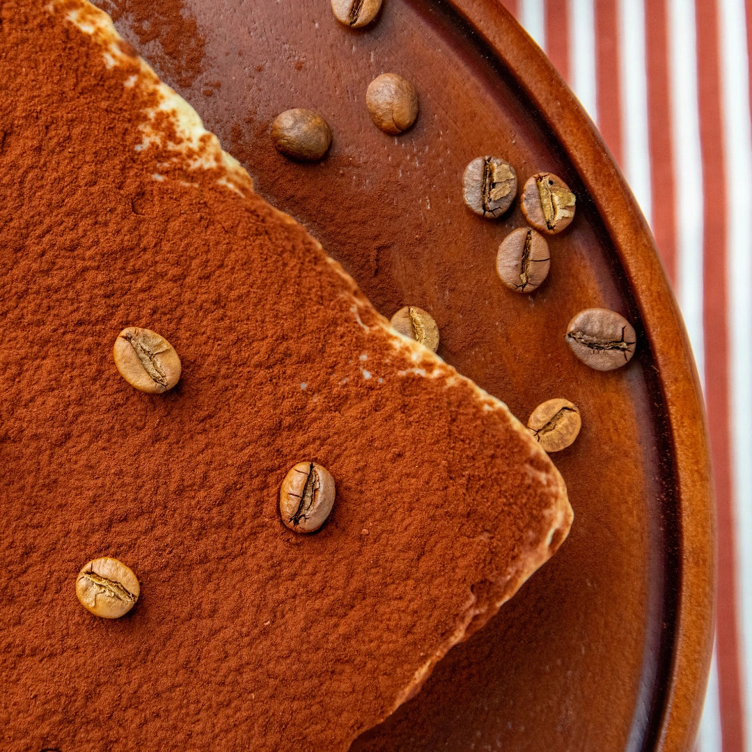 A close-up of the Pick‑Me‑Up tiramisu brownie on a brown plate, topped with cocoa powder and scattered coffee beans, set against a red and white striped cloth