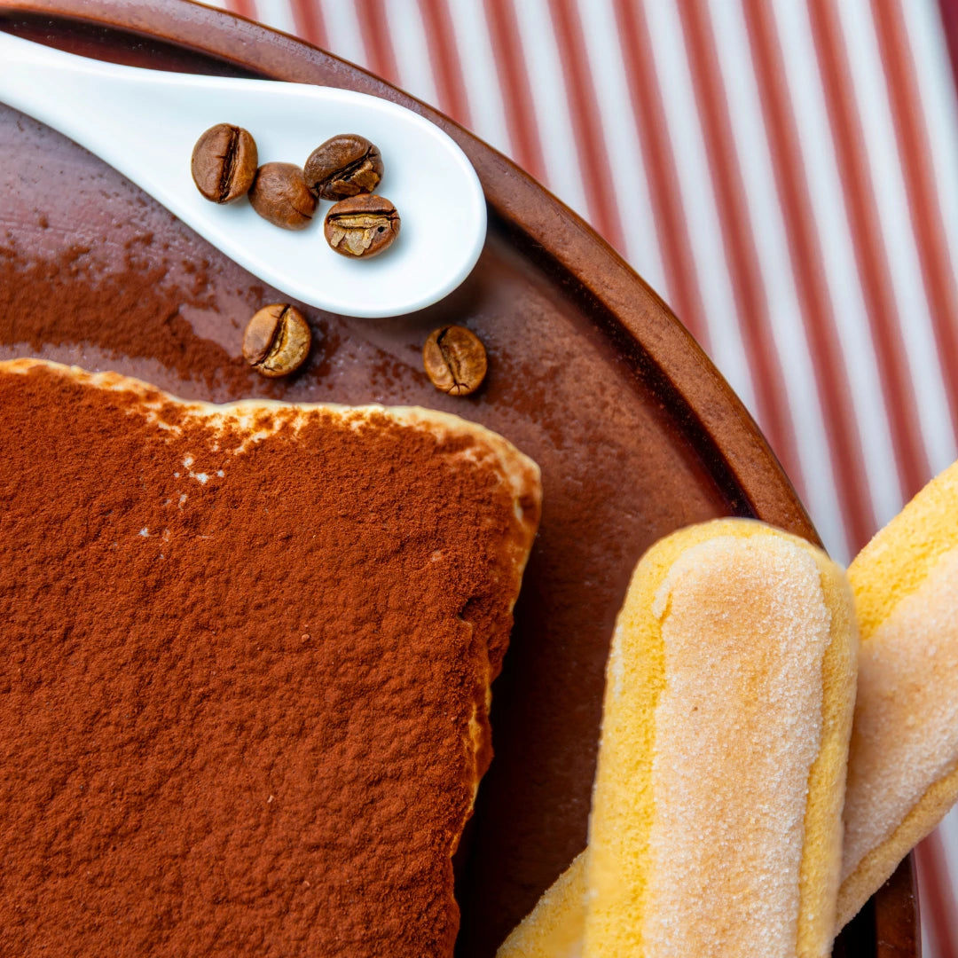 A Pick‑Me‑Up tiramisu brownie slice on a brown plate with a white spoon holding coffee beans and two sugar dusted ladyfingers beside it, styled on a red and white striped background