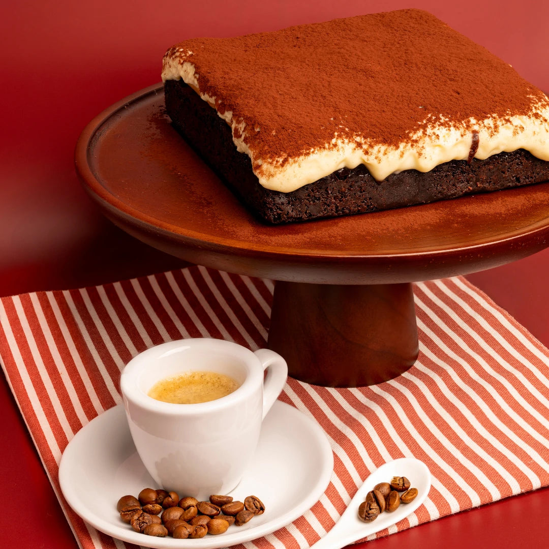 A square Pick‑Me‑Up tiramisu brownie on a wooden cake stand, dusted with cocoa and paired with an espresso cup and coffee beans on a red and white striped cloth.