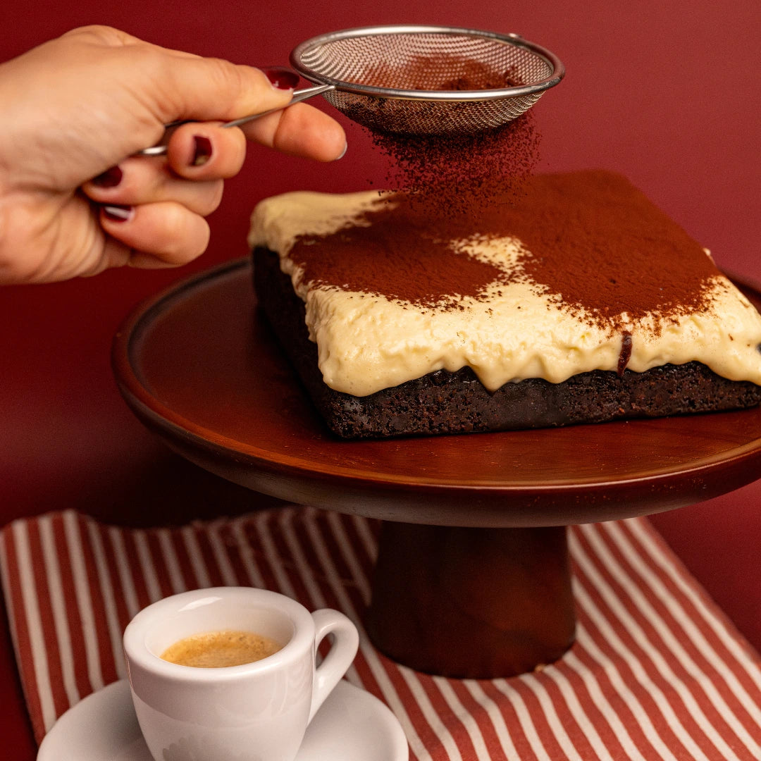 A Pick‑Me‑Up tiramisu brownie being dusted with cocoa powder through a fine mesh strainer, with an espresso cup in the foreground on a red and white striped cloth