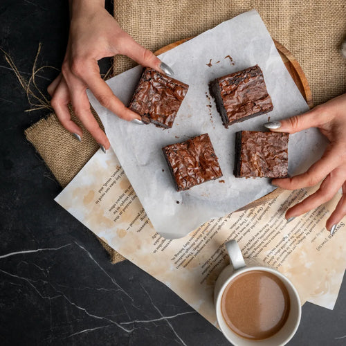 Top-down view of Classic Charm chocolate brownies on parchment paper, with two hands reaching in and a warm coffee mug nearby, creating a cosy dessert moment.