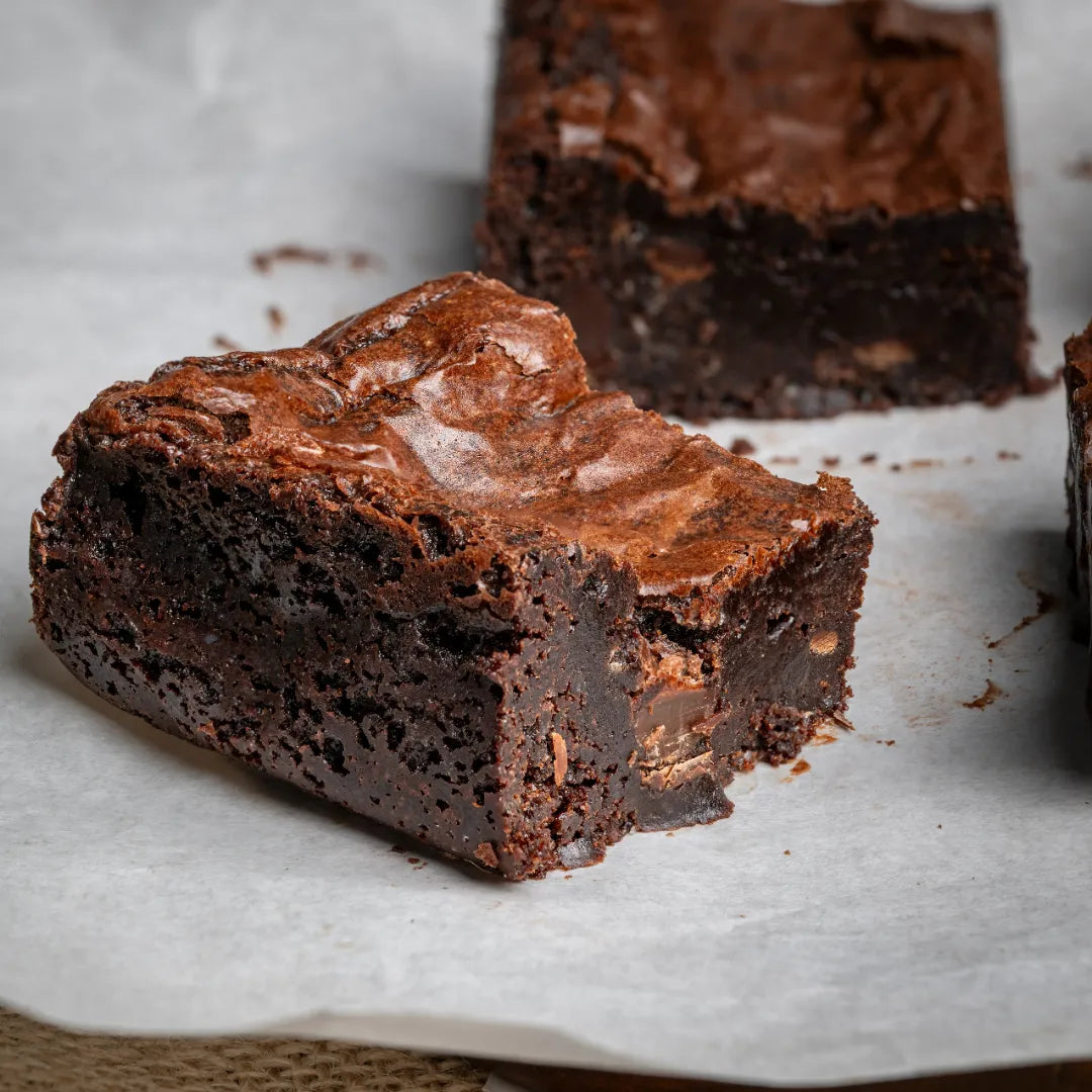 Close-up of a Classic Charm brownie featuring a dense, fudgy interior, cracked shiny crust, and visible chocolate chunks, displayed on parchment for bakery-style presentation.