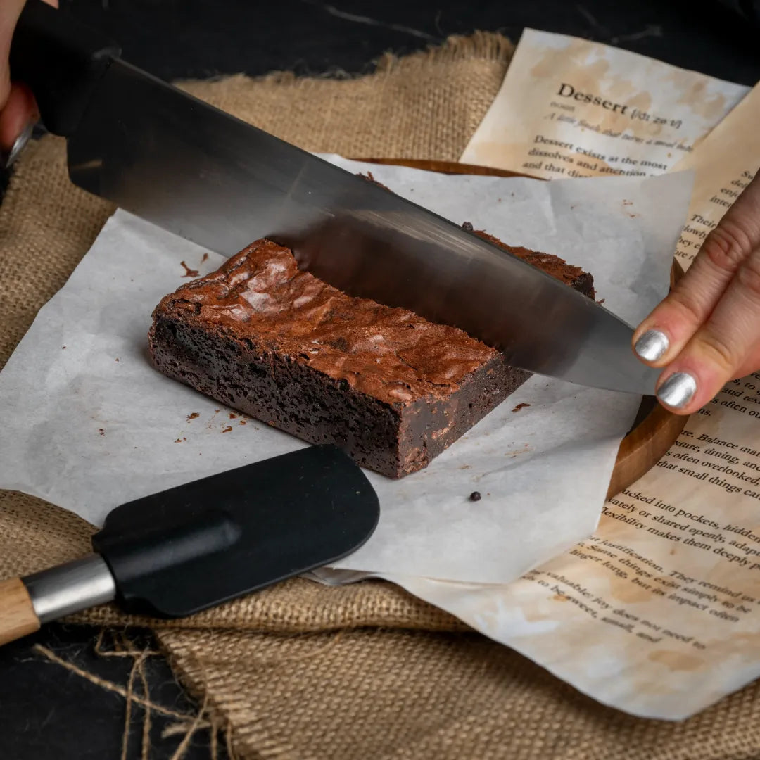 Person slicing a large Classic Charm chocolate brownie on parchment paper with a kitchen knife, surrounded by rustic props and recipe pages for a homemade dessert scene.