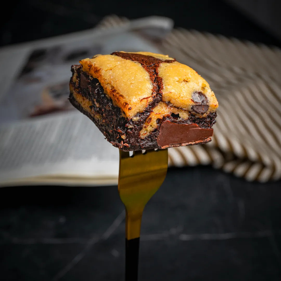 Close-up of a Doughlicious brownie with a cookie‑dough top layer held on a golden fork, highlighting its fudgy chocolate base and soft, chocolate‑chip‑studded cookie dough.