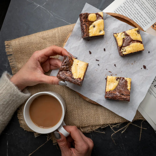 Cosy scene with four Doughlicious cookie‑dough brownies on parchment paper, alongside a hand holding coffee and a marbled brownie for a warm, indulgent moment.