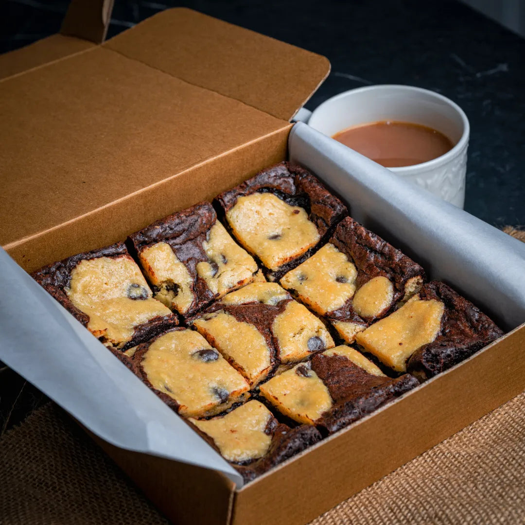 Box of Doughlicious brownies featuring a marbled cookie‑dough topping and chocolate chips, neatly arranged in a parchment‑lined box next to a warm cup of coffee.