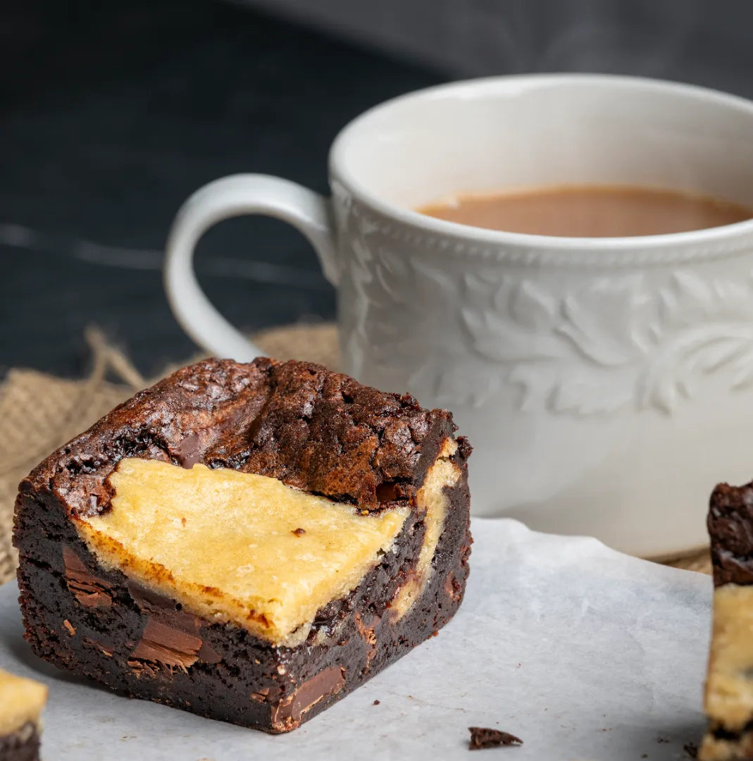 Close-up of a Doughlicious brownie with a fudgy chocolate base and golden cookie‑dough top, served beside a warm drink on parchment paper for a cosy dessert setup.