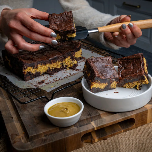 Person lifting a square Dubai Brownie with a spatula, showing its dark chocolate layers and bright yellow centre, surrounded by neatly cut pieces on a cooling rack.