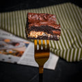 Close-up of a Halva Brownie held on a golden fork, showing its fudgy chocolate layers and creamy helwa tat‑tork filling for a rich, indulgent bite.