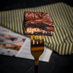 Close-up of a Halva Brownie held on a golden fork, showing its fudgy chocolate layers and creamy helwa tat‑tork filling for a rich, indulgent bite.