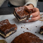 Hand holding a Halva Brownie with a creamy helwa tat‑tork centre, surrounded by more brownies on parchment paper for a cosy dessert scene.