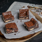 Four Halva Brownies on parchment paper, each with a dark chocolate exterior and a visible helwa tat‑tork cream layer, including one with a bite revealing its texture.
