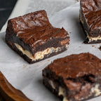 Three thick Halva Brownies on parchment paper, highlighting their fudgy chocolate tops and contrasting helwa tat‑tork filling for a decadent presentation.