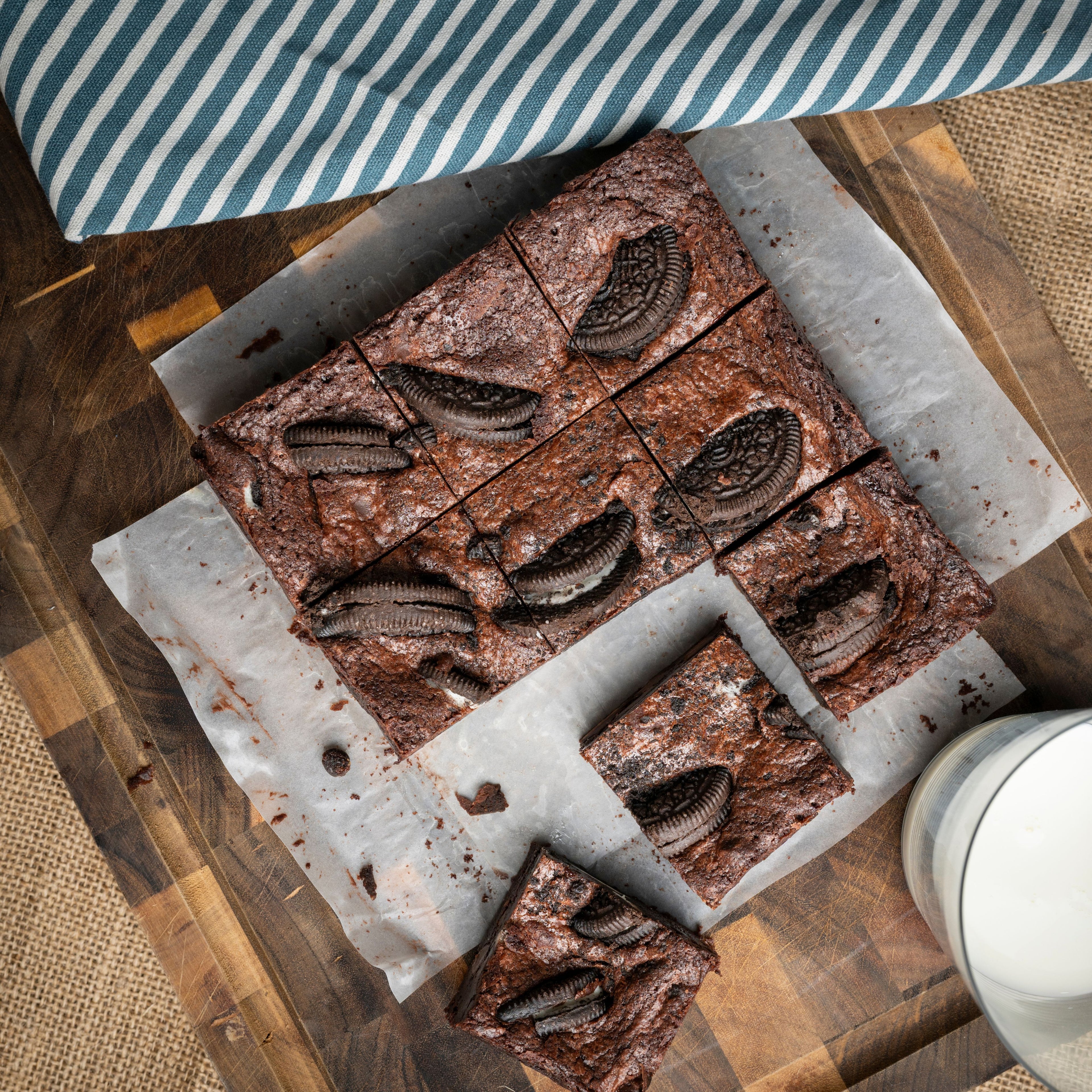Square chocolate brownies topped with Oreo cookie halves, displayed on parchment paper with a glass of milk and striped cloth for a cosy dessert photoshoot aesthetic.