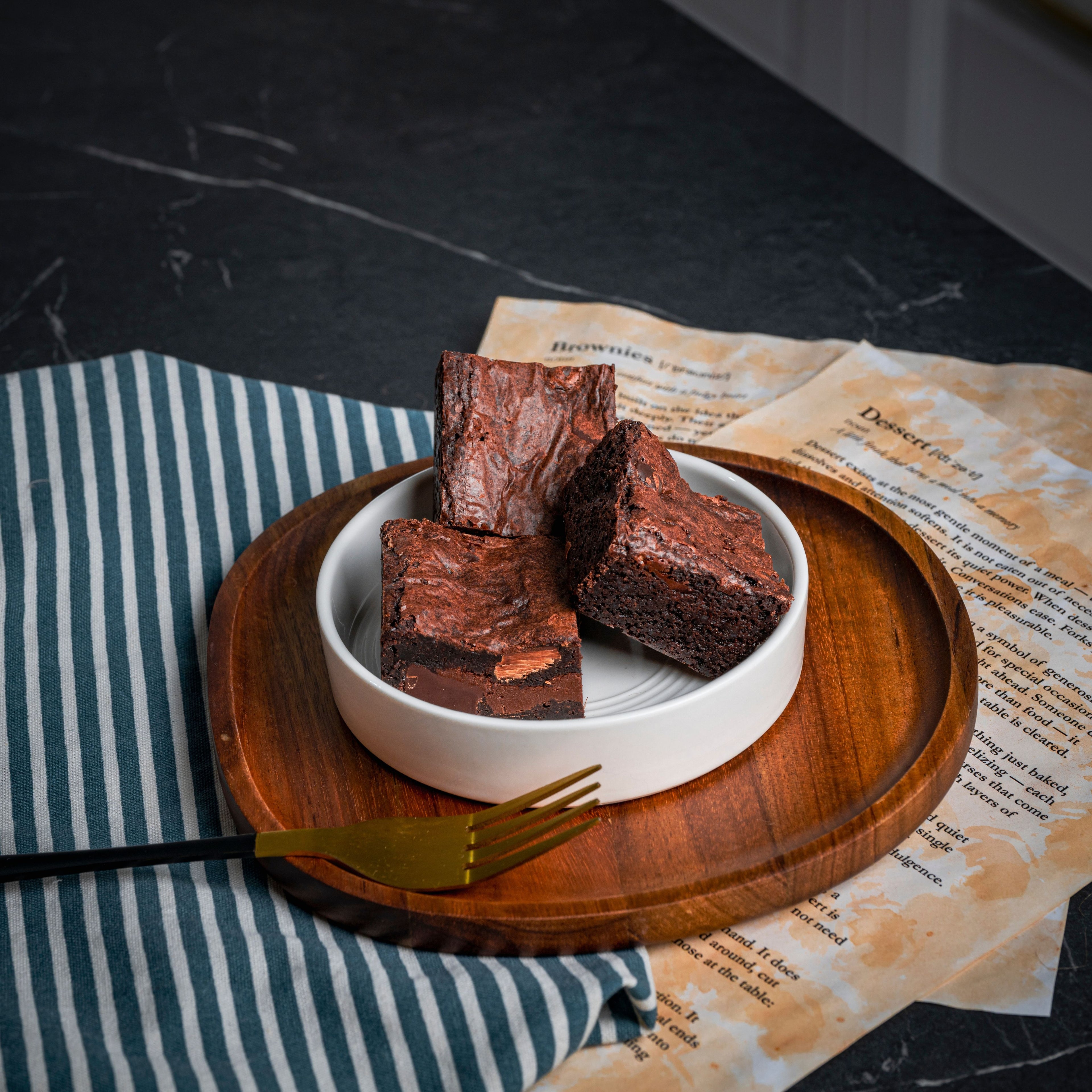 Three fudgy chocolate brownies served in a white ceramic bowl on a wooden tray, styled with a gold fork, striped cloth, and recipe pages in the background for a cosy dessert.