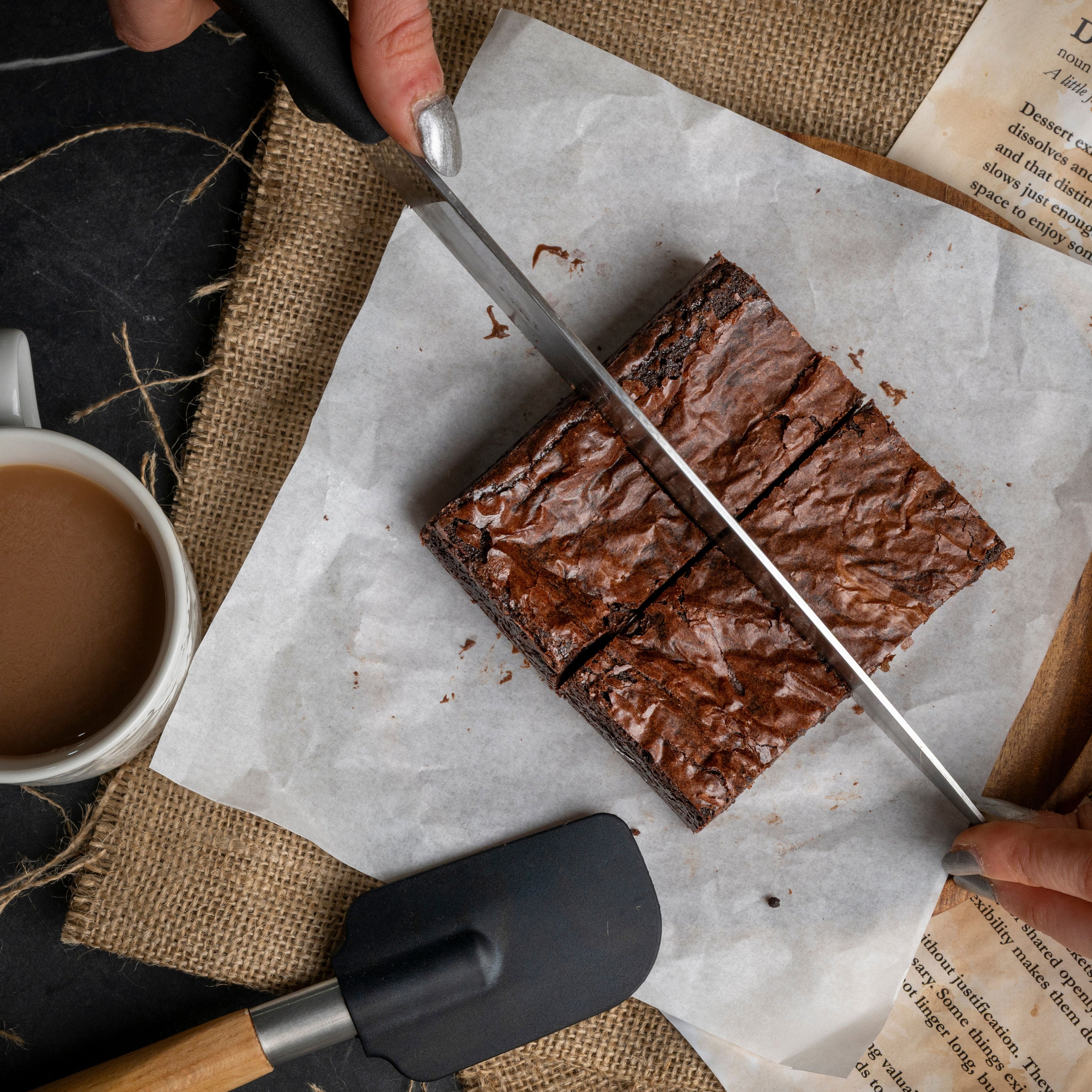Top‑down view of a person slicing a batch of chocolate brownies on parchment paper, styled with a cup of coffee and rustic props for a warm, homemade dessert scene.