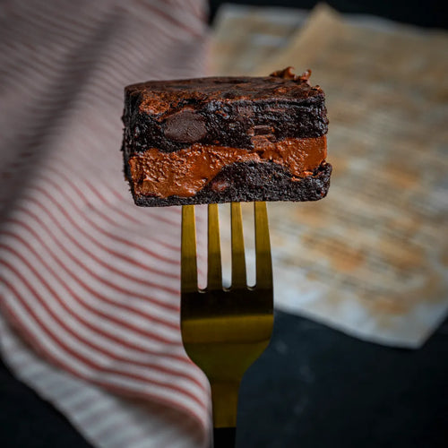 Close-up of a Nuttelluxe brownie held on a golden fork, featuring rich fudgy chocolate layers and a thick Nutella-filled centre for an indulgent bite.