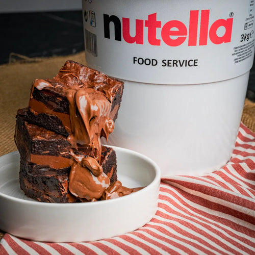 Stack of Nuttelluxe brownies in a white bowl, topped with melting Nutella, with a large Nutella food-service tub in the background for a decadent presentation.