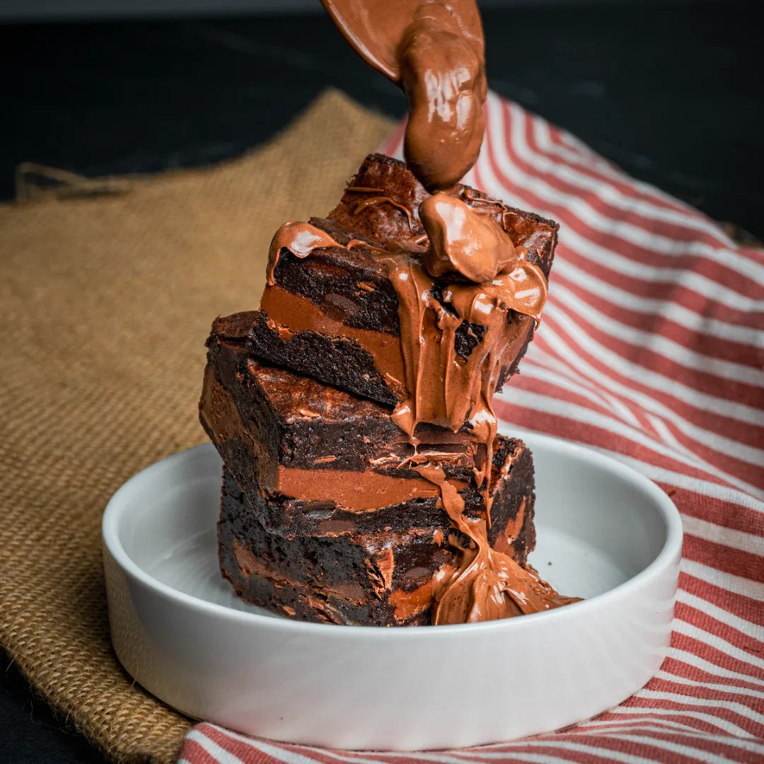 Stacked Nuttelluxe brownies in a white dish with warm melted chocolate being poured over them, creating a rich, Nutella-inspired dessert scene.