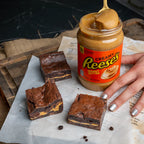 Jar of Reese’s peanut butter beside three Nutty Buddy brownies with a visible peanut butter centre, styled on parchment paper for a rustic dessert scene.