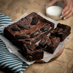 Batch of Oreo Dreams brownies topped with crushed Oreo pieces, with cut squares arranged on parchment paper and a hand holding a glass of milk nearby.