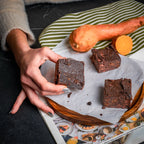 Hand holding a Sweet Surprise brownie beside whole and sliced sweet potatoes, highlighting its flourless, sweet‑potato‑based texture