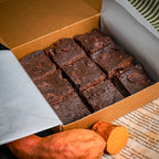 Box of nine Sweet Surprise brownies with whole and sliced sweet potatoes in the foreground, showcasing their naturally fudgy, flourless texture.