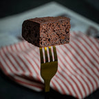 Close-up of a Wine & Dine brownie held on a golden fork, showing its rich dark‑chocolate, cake‑like texture infused with espresso coffee and Merlot wine.