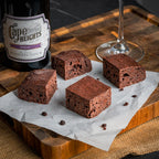 Four Wine & Dine brownies on parchment paper beside a bottle of Merlot and a wine glass, highlighting their cake‑like dark‑chocolate crumb enriched with espresso and wine.