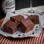 Four Wine & Dine brownies on a wooden plate with a bottle of Merlot in the background, showcasing their cake‑like dark‑chocolate crumb infused with espresso and wine.